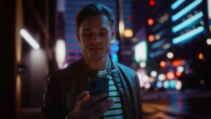 男人使用智能手机走过充满霓虹灯的夜晚城市街道。微笑的时尚男人使用手机，社交媒体，在线购物，在约会应用