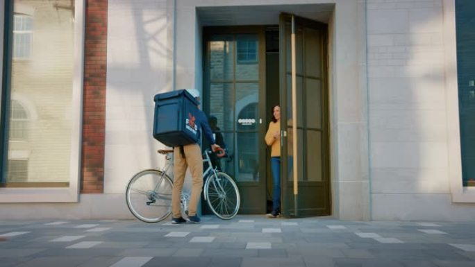 快乐的送餐男子在自行车上穿着保暖背包，向一位美丽的女性顾客提供餐厅订单。快递公司为城市建筑中的漂亮女