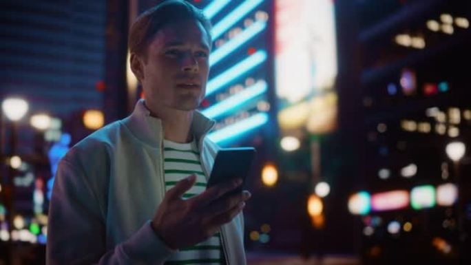 使用智能手机走过充满霓虹灯的夜晚城市街道的男子肖像。微笑的时尚男人使用手机，社交媒体，在线购物，在约