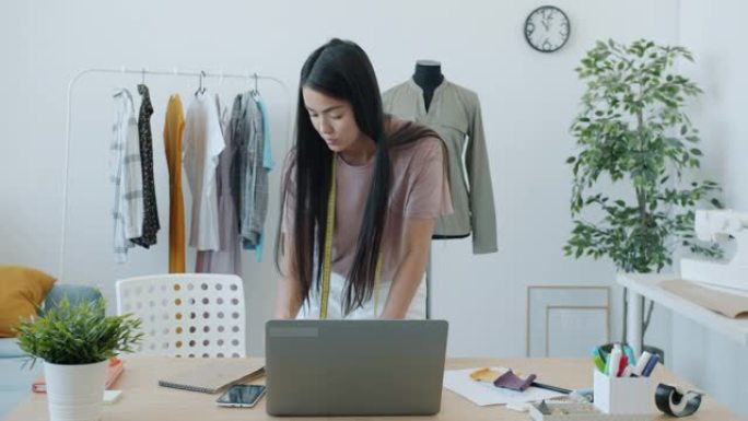 自信的亚洲女裁缝在工作室室内使用笔记本电脑打字和选择材料