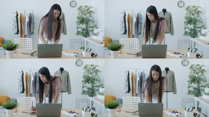 自信的亚洲女裁缝在工作室室内使用笔记本电脑打字和选择材料