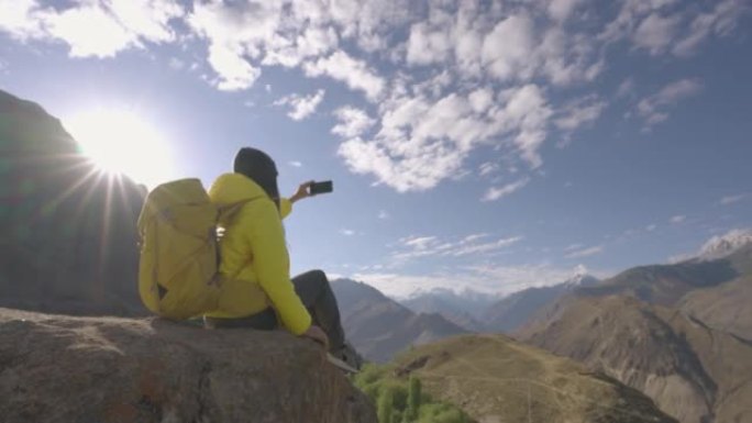 自由旅行者的年轻女性穿着黄色的冬季夹克，背着背包，在早晨的山顶上，用智能手机旅行假期拍摄美丽的景色。