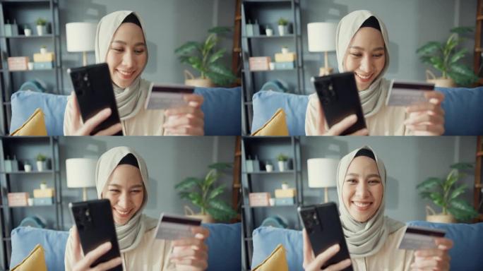 迷人开朗的年轻亚洲伊斯兰穆斯林美女微笑女孩戴着头巾，随意坐在沙发上使用手机在家里舒适的客厅用信用卡享