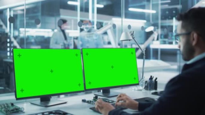 机器人工程师在一家启动工厂的台式计算机上工作，该计算机具有两个绿屏Chromakey显示屏。高科技技