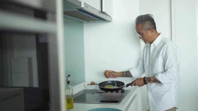 亚洲中国高级男子早上在厨房用炊具打破蛋壳煎蛋准备早餐