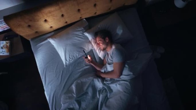 顶景公寓卧室: 英俊的年轻人晚上在床上使用智能手机。微笑的家伙浏览社交媒体，约会应用程序，远程工作软