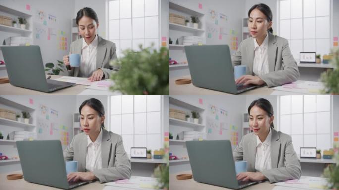 年轻的亚洲开朗的专业女商人财务顾问顾问坐在桌子上，笔记本电脑在现代办公室在线喝咖啡。