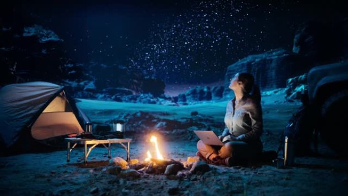 峡谷夜间帐篷露营: 女性旅行者使用坐在篝火旁的笔记本电脑。女性从事数字远程工作，电子购物，电子商务，