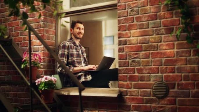 英俊的创意男性坐在他舒适公寓的窗台上，在笔记本电脑上工作。专业专家在家远程工作，听鸟儿唱歌。