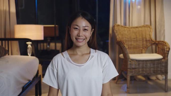 愉快的年轻亚洲女士晚上在家客厅里用手机看着摄像头进行视频直播。