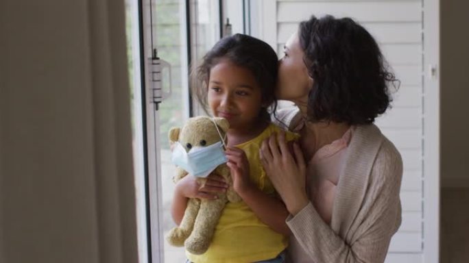 西班牙裔母女拥抱在窗户上，戴着口罩的泰迪熊