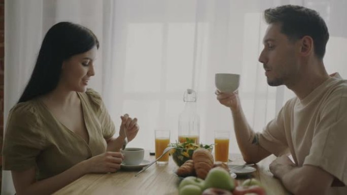 幸福的年轻夫妇吃早餐喝咖啡，坐在家里厨房的桌子旁聊天