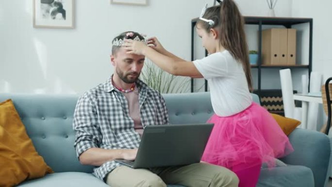 可爱的孩子用bijouterie装饰父亲，而男人正在家里用笔记本电脑工作