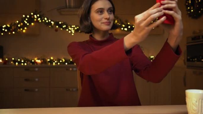 一个年轻漂亮的女人正在她的红色智能手机上自拍，坐在一张拿着蜡烛的桌子旁，在圣诞灯下的厨房里