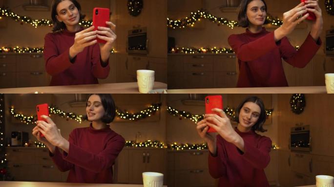 一个年轻漂亮的女人正在她的红色智能手机上自拍，坐在一张拿着蜡烛的桌子旁，在圣诞灯下的厨房里