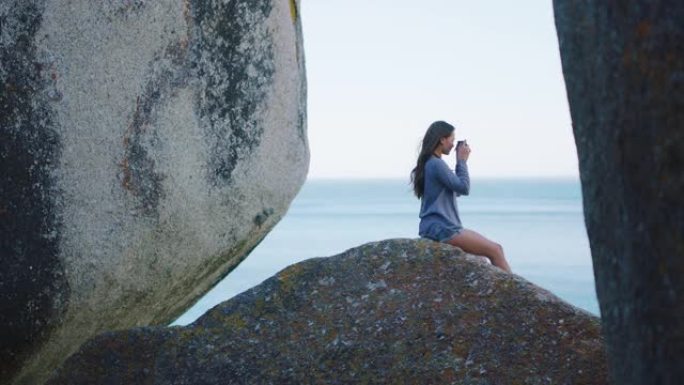 摄影师，海滩和和平与一个女人在大自然中，坐在岩石巨石上欣赏地平线的海景。水上，旅行和自由与一名女性在