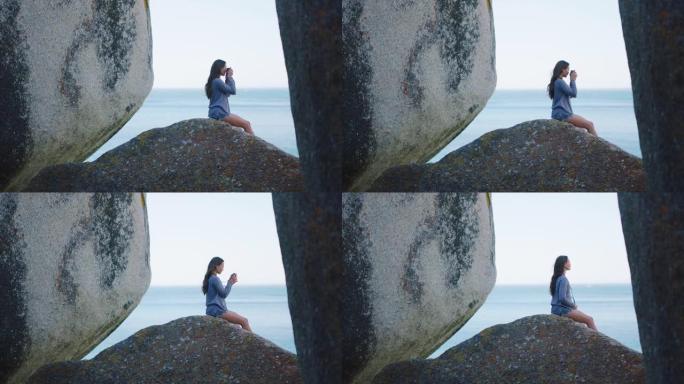 摄影师，海滩和和平与一个女人在大自然中，坐在岩石巨石上欣赏地平线的海景。水上，旅行和自由与一名女性在