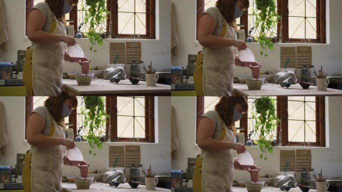 戴着口罩的女性高加索波特在陶器工作室的锅上倒油漆
