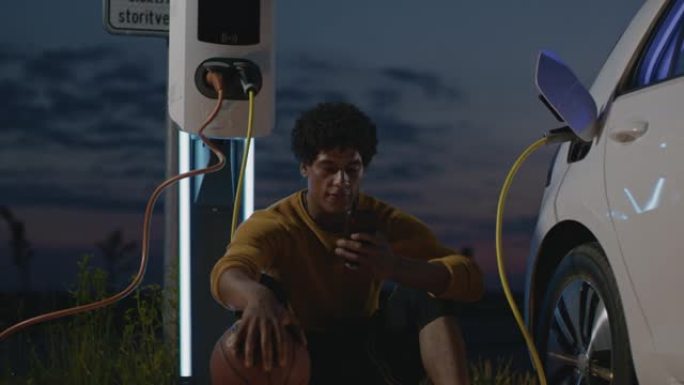 SLO MO年轻的篮球运动员晚上在停车场用智能手机充电时使用智能手机