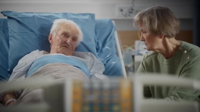 医院病房: 老人躺在床上休息，他有爱心的美丽妻子来访，支持他坐在旁边，手牵着手，幸福的夫妻交谈。老人
