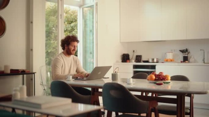英俊的成年男子，姜黄色卷发，使用笔记本电脑，坐在公寓的客厅。男性喝浓缩咖啡，在家工作，网上购物，看视