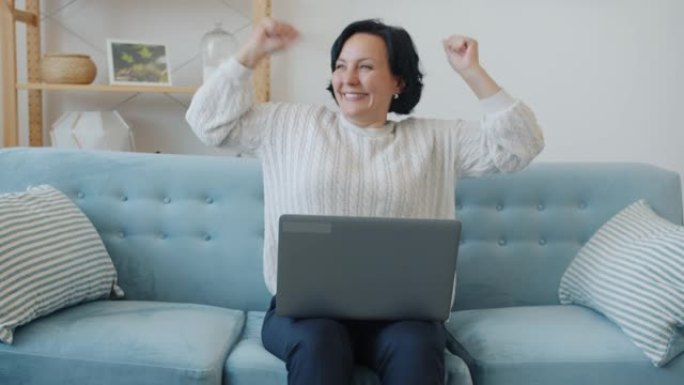 兴奋的女士慢动作用笔记本电脑举起手臂在公寓里享受好消息