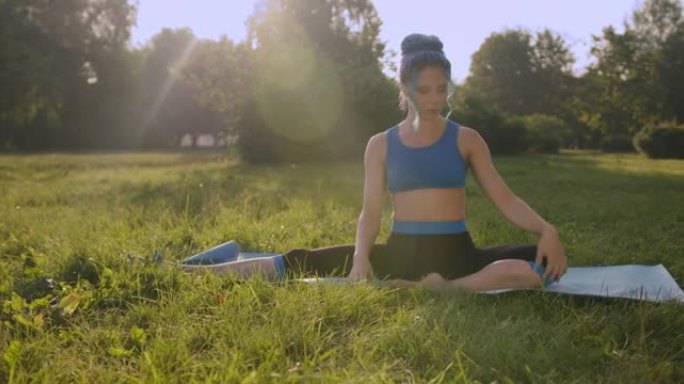 迷人的年轻女子，蓝色的头发，穿着蓝色的瑜伽服，在公园的草地上做瑜伽，在美丽的阳光下左右伸展到直腿，一