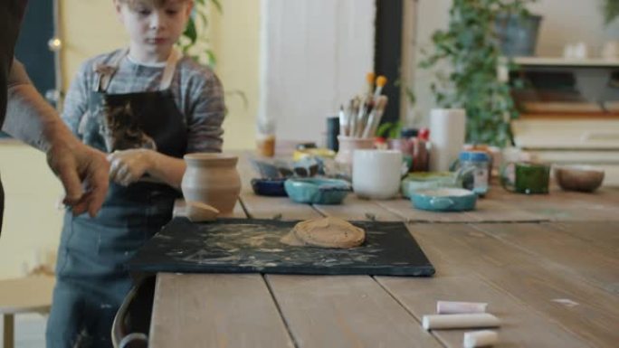 成年男子的手在陶器工作室里用粘土工作，而孩子在围裙里看着