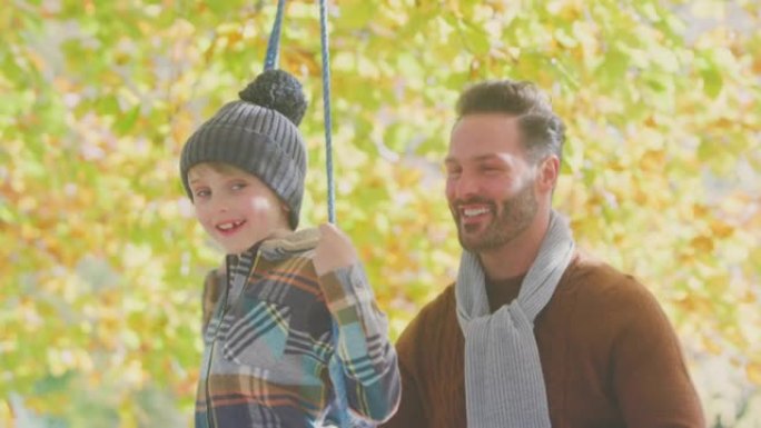 父亲推着儿子在秋季花园的绳索秋千上玩得开心