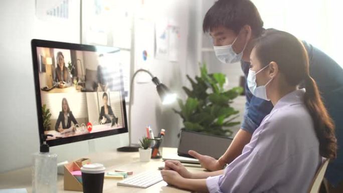 戴着口罩的高加索商人在办公室的计算机屏幕上与她的亚洲男同事，商人一起工作的视频会议