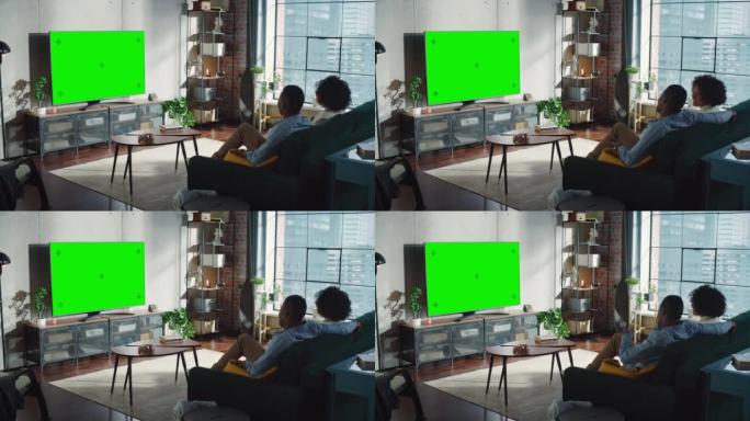 黑人夫妇坐在家里的客厅里，看绿色色度键屏幕电视，在沙发上放松。周末娱乐概念
