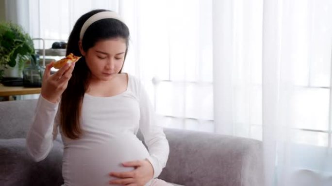 亚洲妇女在家里的沙发上在怀孕期间吃披萨不健康的食物。垃圾食品