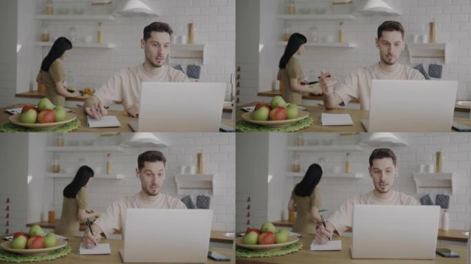 在家工作的年轻人用笔记本电脑进行在线视频通话，而妻子在后台做午餐
