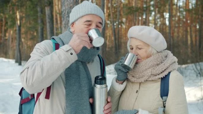 快乐的高级夫妇碰杯喝热水瓶里的热茶，在冬季公园户外聊天