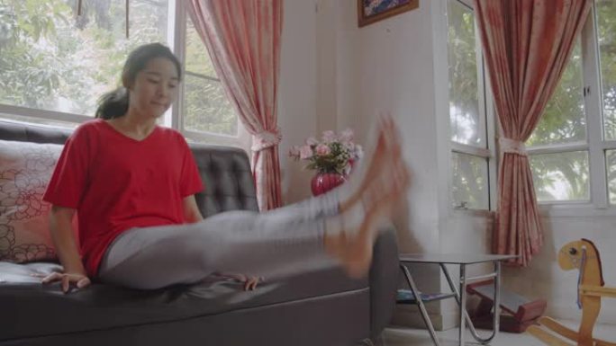 迷人的亚洲少女坐在沙发上，在客厅做抬腿运动。女孩在家时用体重锻炼。