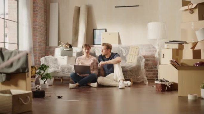 快乐的年轻房主: 坐在新买公寓地板上的夫妇使用笔记本电脑。在网上购物时做团队合作击掌。抵押贷款，房地