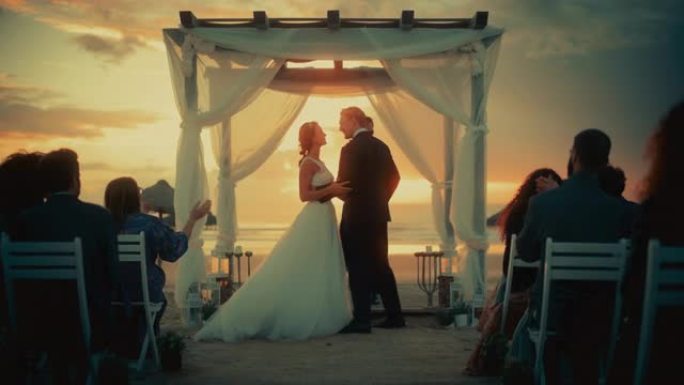 美丽的新娘和新郎在日落时在海洋海滩举行的户外婚礼上。浪漫情侣结婚、交换戒指、亲吻和与朋友分享庆祝活动
