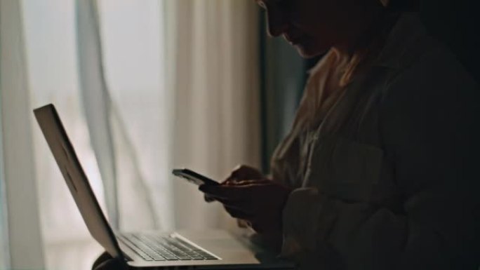 特写女人使用她的智能手机和笔记本电脑，坐在酒店房间的床上，背景是窗户和窗帘，慢动作