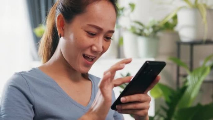 特写年轻的亚洲女性穿休闲网站在沙发上使用智能手机感觉令人兴奋的滚动屏幕在线购物在客厅室内植物在家。