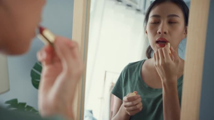 穿着休闲布的美丽的亚洲女士早上在家里的卧室里，在镜子前把口红涂在嘴唇上，然后出去约会。