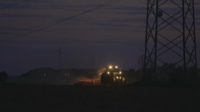 SLO MO拖拉机在夜间在电塔架下犁地