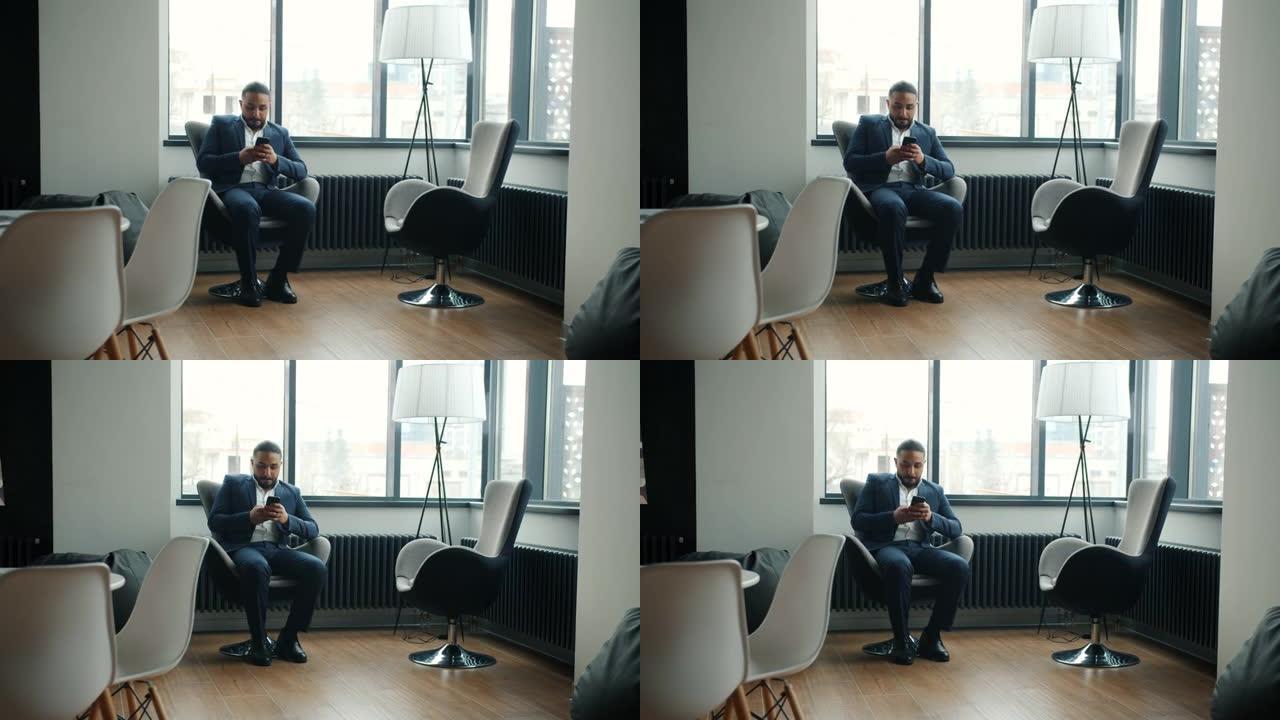 穿着西装的中东男人在现代办公室用智能手机坐在扶手椅上