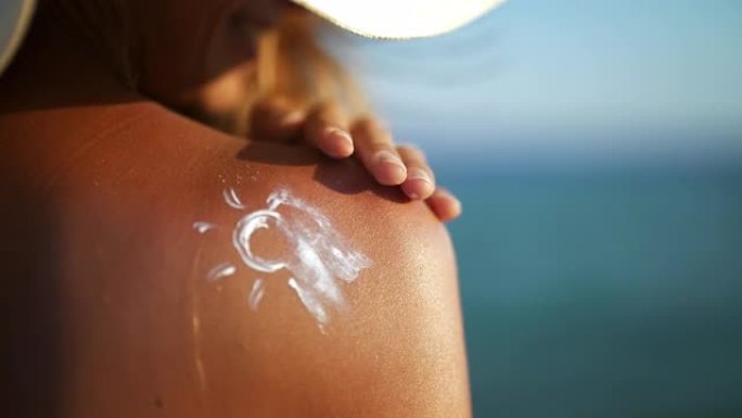 带着草帽的快乐微笑的年轻女子的特写镜头正在肩膀上涂抹防晒霜或晒黑乳液，以在海边的海滩上护理她的皮肤