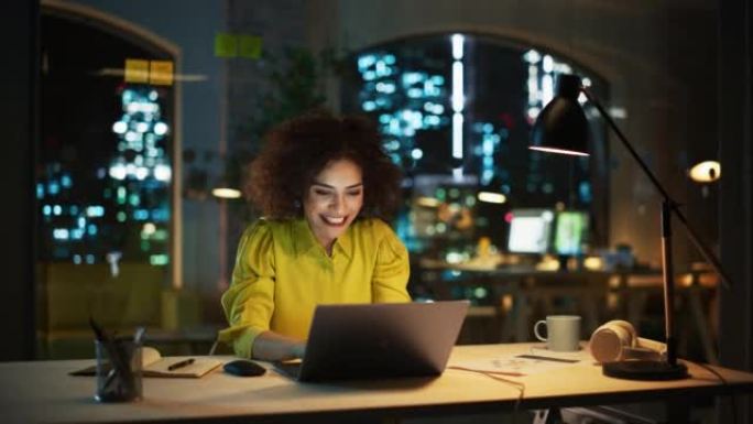 成功的阿拉伯女性坐在创意机构，晚上在笔记本电脑上工作。经理庆祝令人印象深刻的销售业绩。兴奋的专家对她