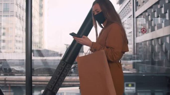 侧视图年轻漂亮的女商人戴着口罩在商场橱窗里用手机办公室应用带着购物袋散步。