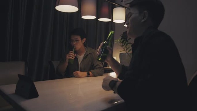 两名亚洲成年男子在家时通过在线视频通话虚拟聚会与朋友一起喝啤酒