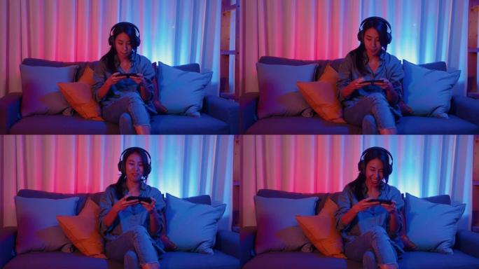 快乐亚洲女孩游戏玩家戴着耳机比赛在线视频游戏与智能手机兴奋的谈话与朋友坐在沙发上在彩色霓虹灯在家里的