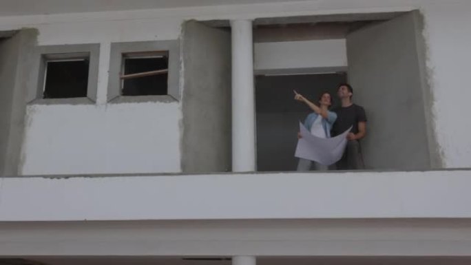 拉丁美洲成年幸福夫妇站在正在建造的房屋的二楼，拿着蓝图，指着远处