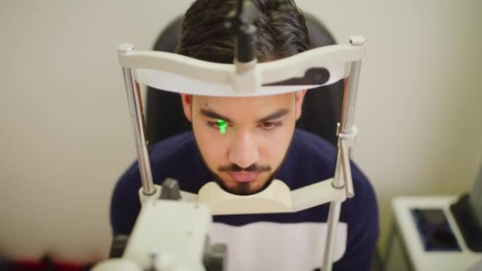 视光师的男性患者在每年的约会中进行视网膜眼睛扫描以检查其视力。眼科医生的拉丁裔男子从上方检查视力问题