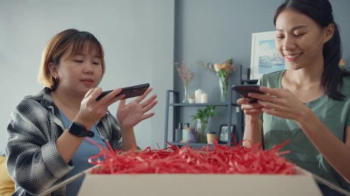 快乐亚洲女士朋友从在线市场打开纸板箱包装拍照以进行反馈评论，以在家中客厅购买网站社交媒体。